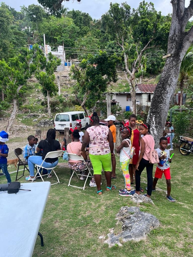 Children and adults gather outdoors in a lush, green space, engaging socially around chairs and a table, creating a community atmosphere.