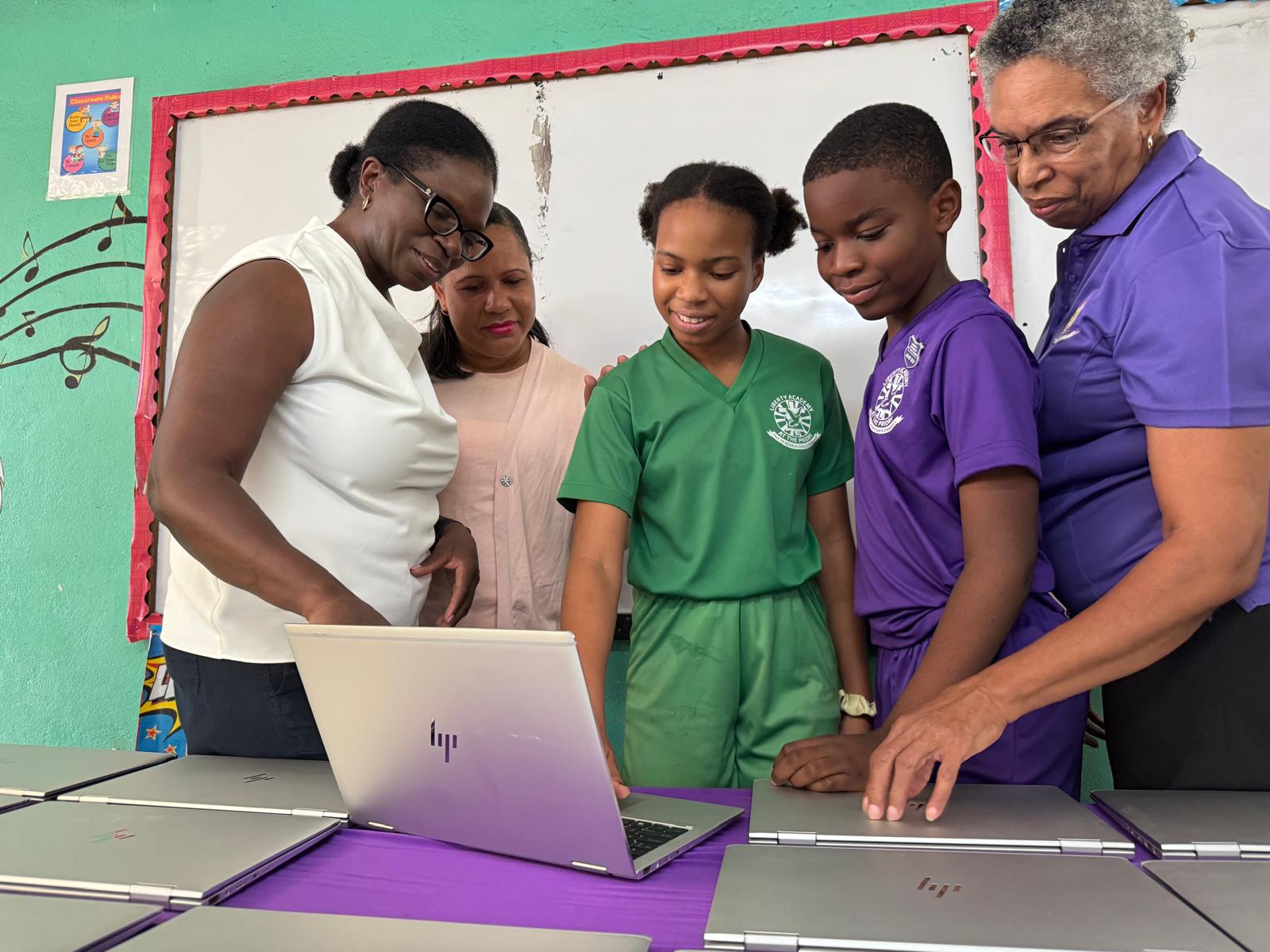 Students and educators gather around a laptop, engaging in a collaborative learning activity with multiple laptops on a purple table.